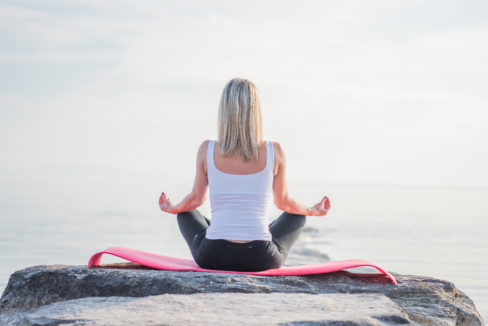 Woman sitting cross-legged and meditating on a rock by the water